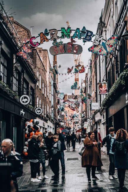 A bustling street scene on Carnaby Street in Soho, featuring a narrow pedestrian lane lined with retail shops and boutiques. The street is decorated with colorful butterflies and festive signs hanging above, creating a vibrant atmosphere. The pavement appears clean and well-maintained, with visitors strolling, some engaged in conversation and others window shopping. The shopfronts display modern signage and window displays, while overhead lighting and street decorations add to the lively ambiance. Overcast skies cast a neutral light over the scene, and the overall cleanliness and well-kept appearance suggest that Soho Carpet Cleaning provides thorough surface cleaning and maintenance services to keep commercial areas tidy and welcoming.