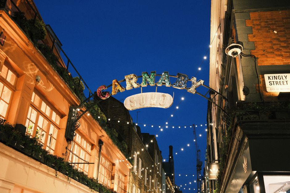 A bustling street scene on Carnaby Street in Soho, featuring a narrow pedestrian lane lined with retail shops and boutiques. The street is decorated with colorful butterflies and festive signs hanging above, creating a vibrant atmosphere. The pavement appears clean and well-maintained, with visitors strolling, some engaged in conversation and others window shopping. The shopfronts display modern signage and window displays, while overhead lighting and street decorations add to the lively ambiance. Overcast skies cast a neutral light over the scene, and the overall cleanliness and well-kept appearance suggest that Soho Carpet Cleaning provides thorough surface cleaning and maintenance services to keep commercial areas tidy and welcoming.
