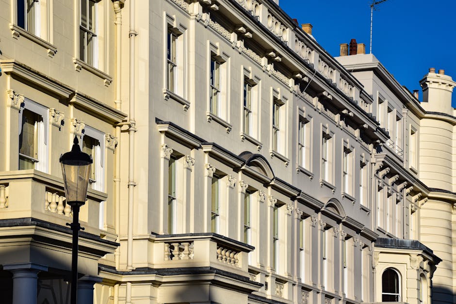 A close-up view of a historic multi-story building with a cream-colored facade in Soho, featuring detailed architectural elements such as ornate window frames, decorative cornices, and small balconies with balustrades. The building has several tall, rectangular windows with white trim, some partially open, and is illuminated by natural sunlight against a clear blue sky. In the foreground, there is a black metal street lamp with a traditional design. The image emphasizes the building's clean exterior surface, with no visible dirt or damage, illustrating the importance of regular surface cleaning and maintenance as promoted by Soho Carpet Cleaning in compliance with Westminster Council rules for disposing cleaning waste in Soho.