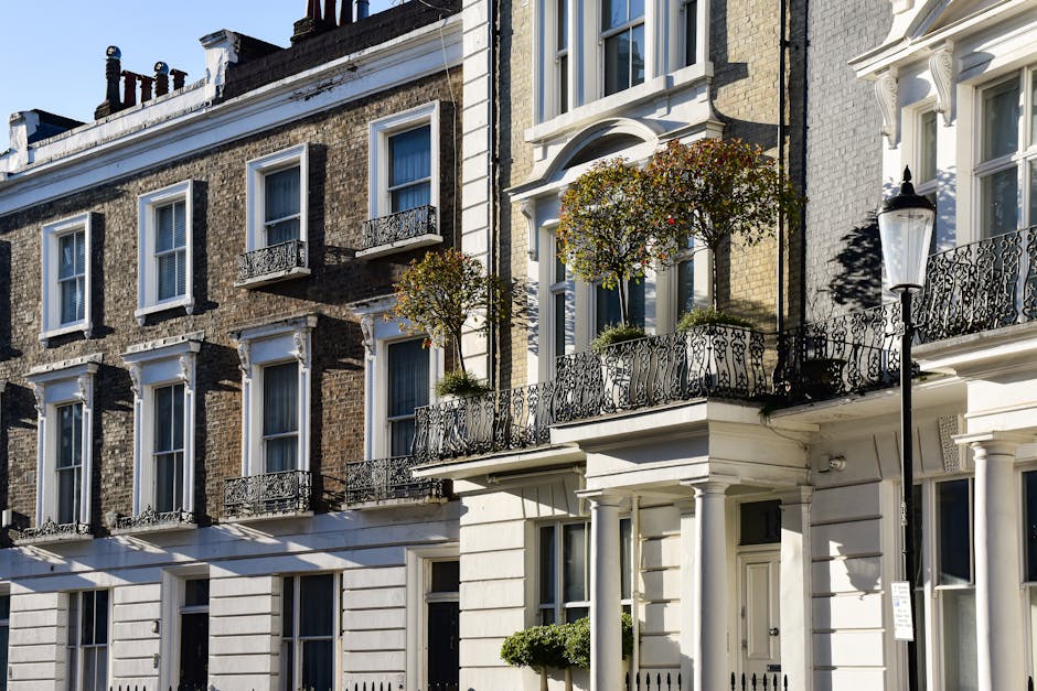 A row of multi-story residential buildings with brick facades and white-painted exterior walls, featuring large windows with white frames and decorative wrought iron balconies. Some balconies have small potted plants and trees, adding greenery to the scene. A traditional black street lamp is positioned in front of the buildings, casting shadows on the facade. The scene is illuminated by bright daylight, highlighting the clean and well-maintained surfaces, including smooth brickwork, polished window sills, and the freshly painted white sections of the building. The overall appearance reflects a tidy, urban residential area consistent with Soho's architecture, maintained with professional cleaning and upkeep by Soho Carpet Cleaning.