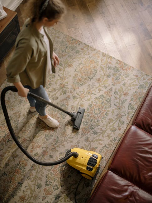 A person using a yellow vacuum cleaner to deep clean a patterned area rug in a living room, with wooden flooring visible around the rug and a brown leather sofa nearby. The individual is wearing casual clothing and white shoes, focusing on surface cleaning to maintain hygiene and cleanliness, with soft lighting illuminating the space. Soho Carpet Cleaning offers professional domestic cleaning services, including carpet and surface sanitisation, as seen in this scene.